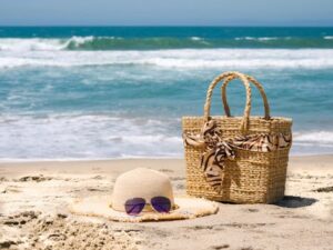 Straw hat, beach bag, and sunglasses on sandy beach with ocean waves.